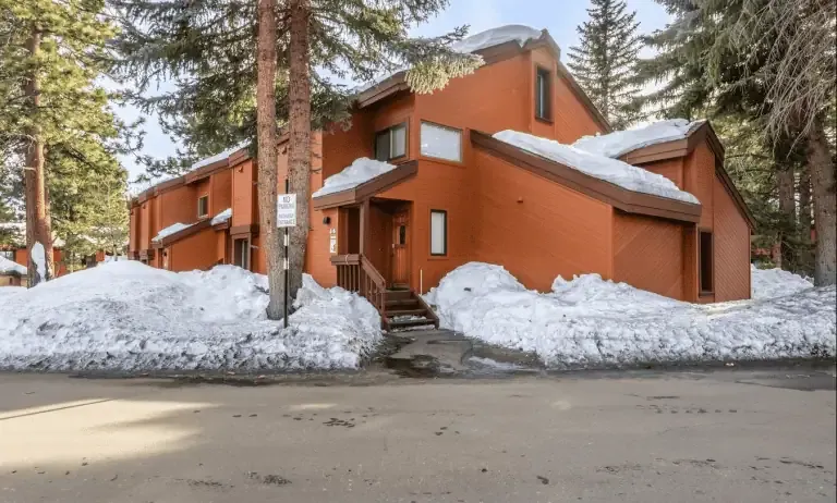 A brown two-story townhouse with a garage and snow piled around the building, surrounded by tall evergreen trees on a clear day.