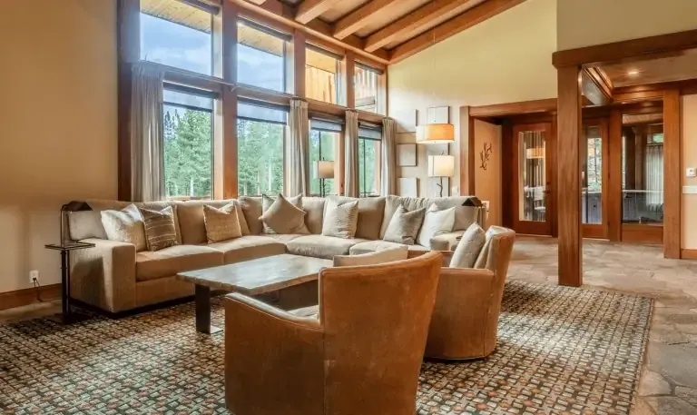 Spacious living room with large windows, beige sectional sofa, two brown leather chairs, wooden coffee table, patterned rug, and high wood-beamed ceiling.