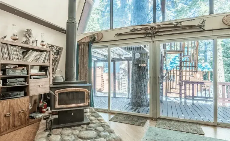 A wood stove sits on a stone platform beside sliding glass doors, which open to a deck with a spiral staircase and a view of trees outside. Shelves with records and audio equipment are on the left.