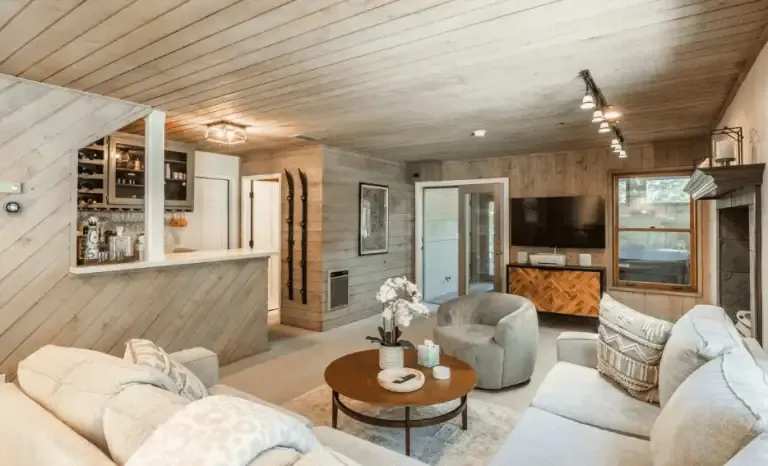 A cozy living room with wood-paneled walls and ceiling, a round coffee table, upholstered seating, a wall-mounted TV, a fireplace, and a small bar area with shelves and wine storage.