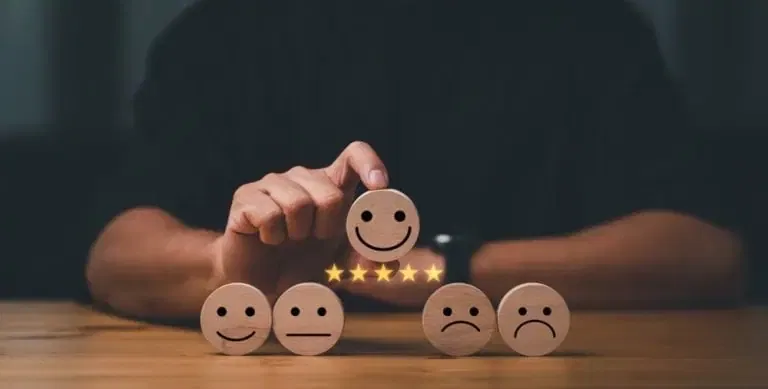 A hand selects a wooden block with a smiling face above five stars, surrounded by blocks with neutral and sad faces, representing customer satisfaction ratings.