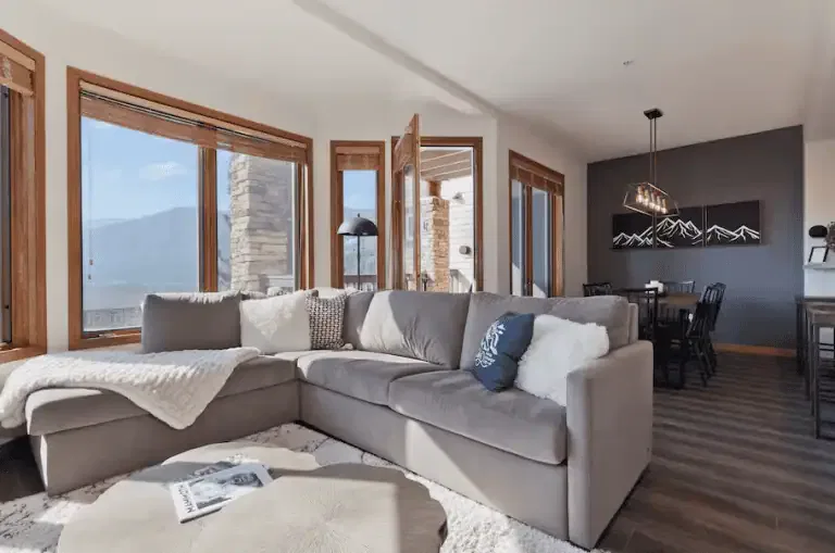 Bright living room with a gray sectional sofa, throw pillows, and blanket, next to large windows; dining area and mountain artwork visible in the background.